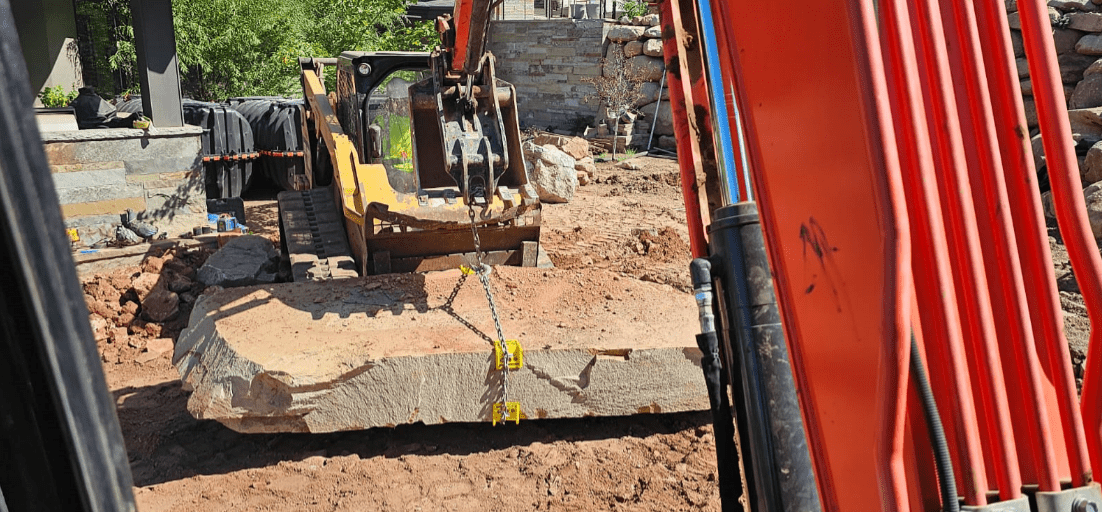 Excavator lifting a large stone slab at a landscaping site in Salt Lake City.