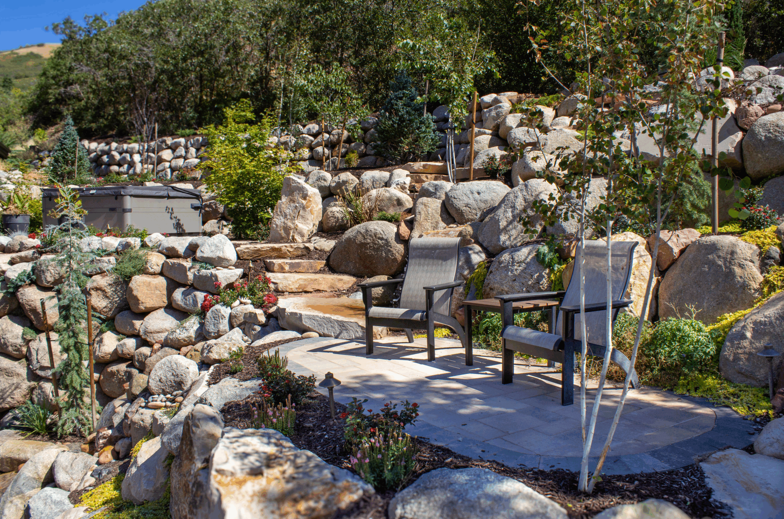 A terraced backyard in Salt Lake City featuring natural stone retaining walls, colorful native plants, and a cozy seating area with two modern chairs—perfectly blending innovative landscape design with the surrounding hillside.