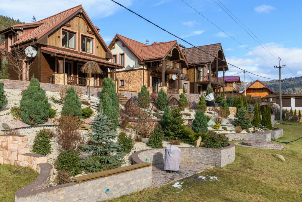 A row of modern mountain-style houses with terracotta roof tiles and wood-accented facades, situated on a landscaped hillside. The foreground features terraced retaining walls, neatly arranged conifer trees, ornamental shrubs, and decorative rock mulch. The homes are surrounded by well-maintained gardens and mountain scenery under a blue sky.