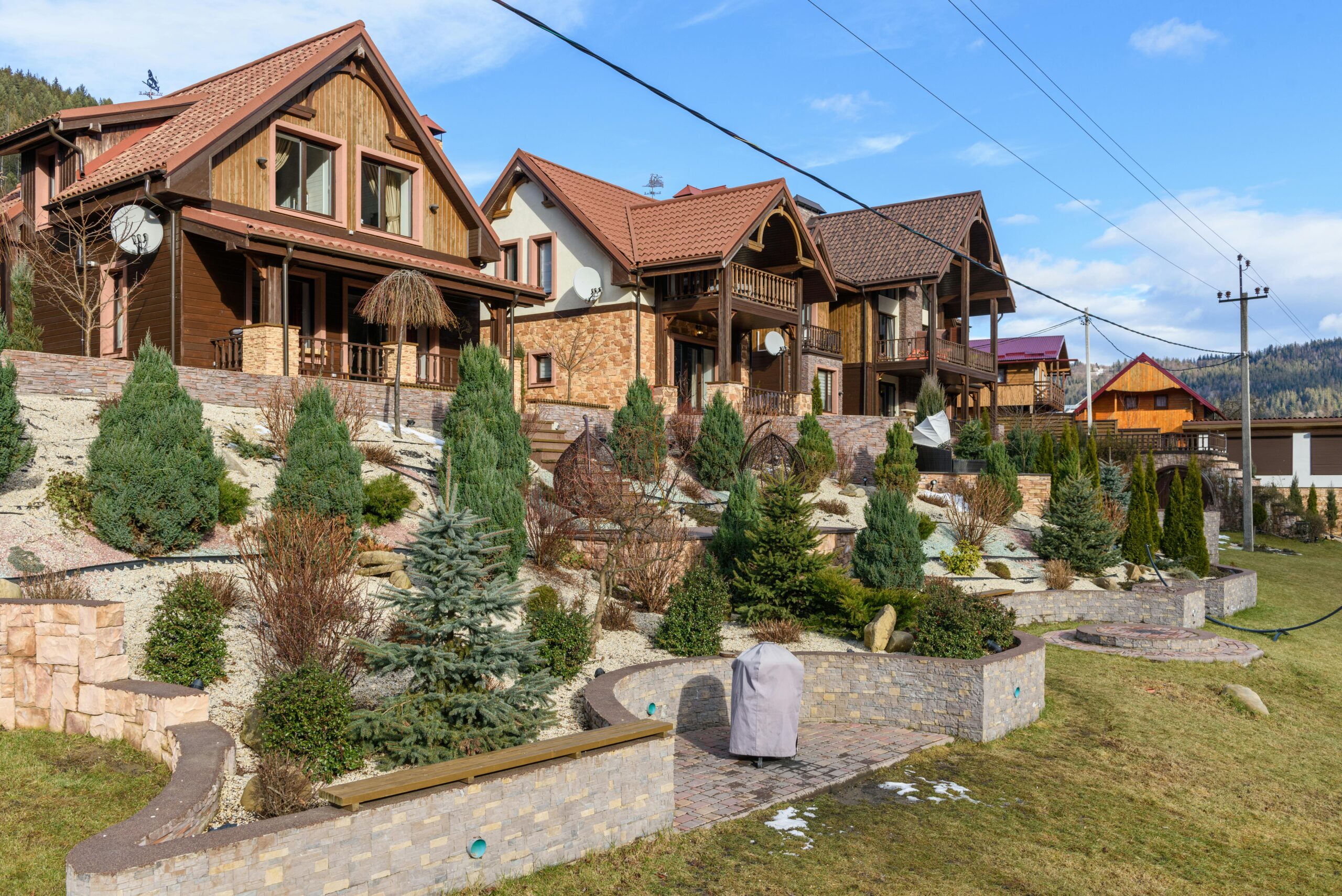 A row of modern mountain-style houses with terracotta roof tiles and wood-accented facades, situated on a landscaped hillside. The foreground features terraced retaining walls, neatly arranged conifer trees, ornamental shrubs, and decorative rock mulch. The homes are surrounded by well-maintained gardens and mountain scenery under a blue sky.