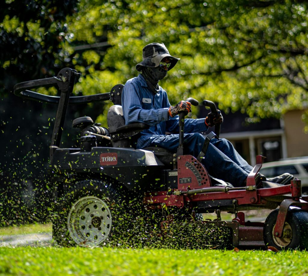 A professional landscaper operating a Toro zero-turn lawn mower on a bright sunny day. The worker is fully covered with protective clothing, including a wide-brimmed hat, gloves, and face covering, with grass clippings visibly flying through the air. The background features lush green trees and a residential area.
