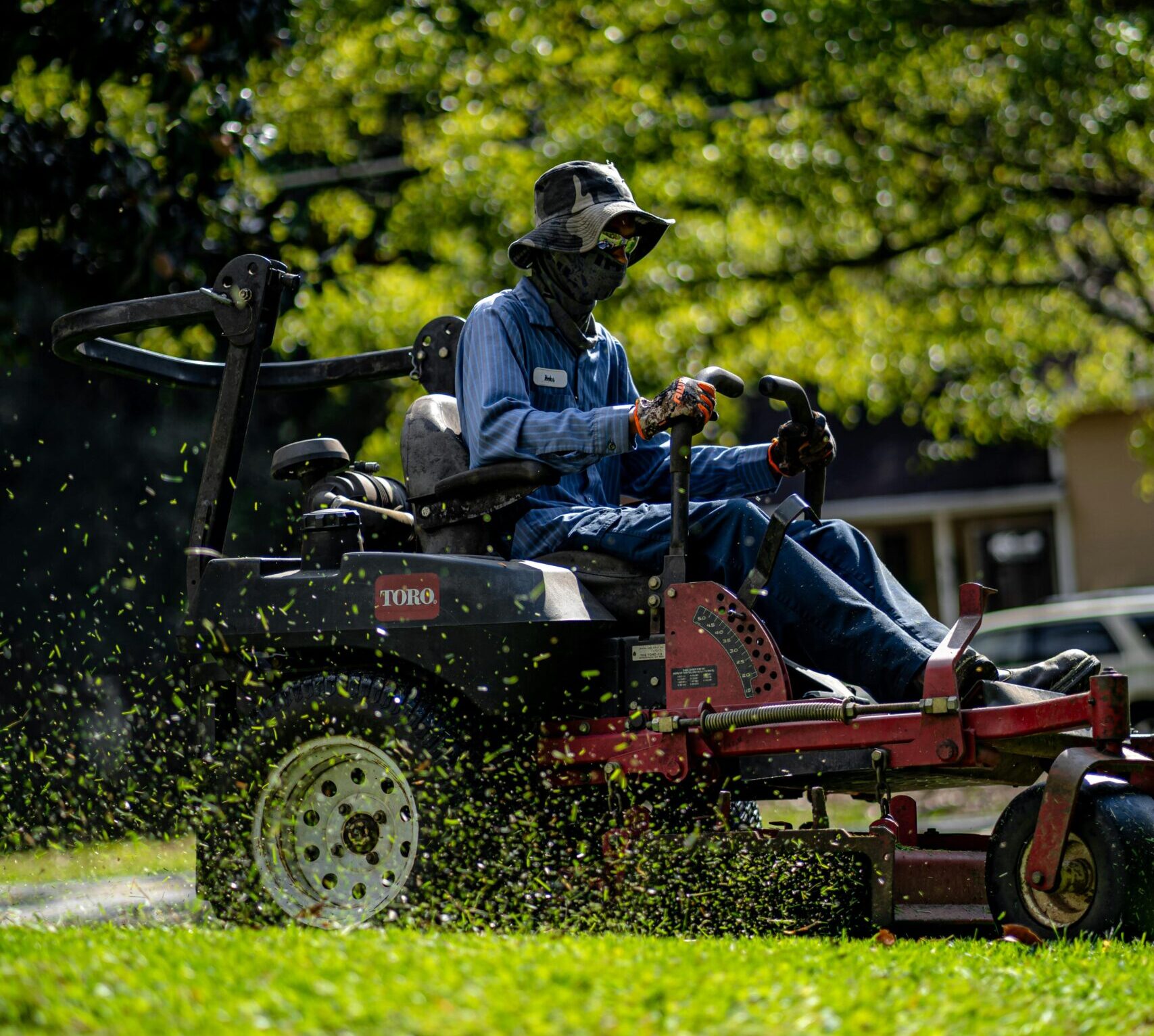 A professional landscaper operating a Toro zero-turn lawn mower on a bright sunny day. The worker is fully covered with protective clothing, including a wide-brimmed hat, gloves, and face covering, with grass clippings visibly flying through the air. The background features lush green trees and a residential area.