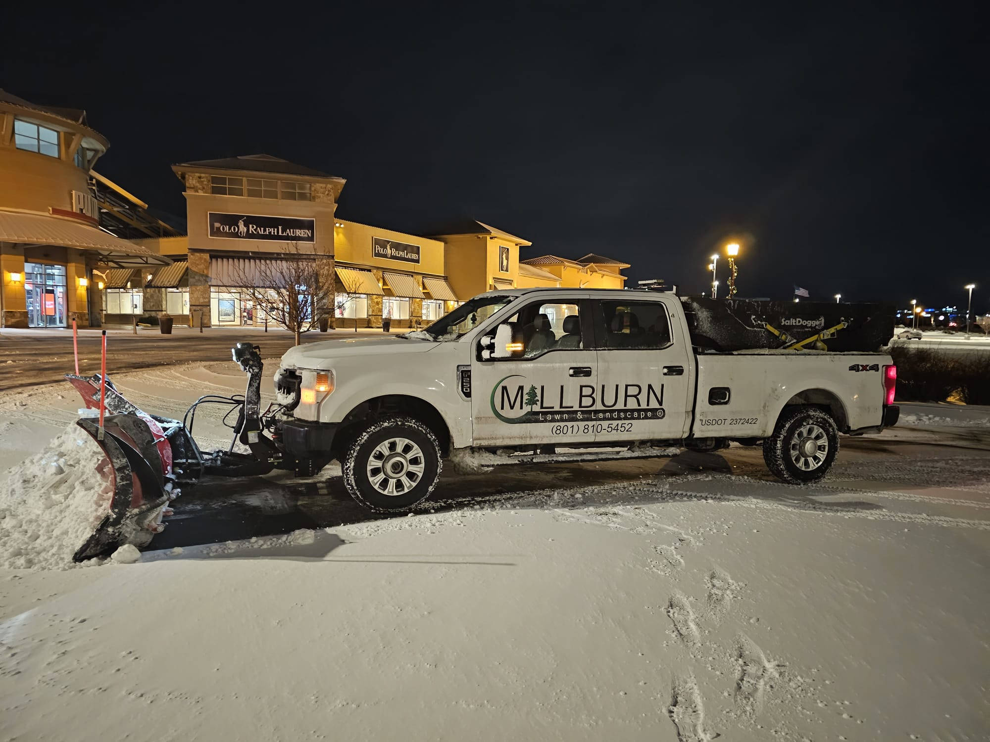 A Millburn Lawn & Landscape snow plow truck clearing snow in front of Polo Ralph Lauren store at night in Utah.