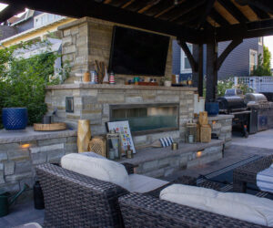 Covered backyard pergola with stone outdoor fireplace, mounted TV, seating area, and built-in grill kitchen