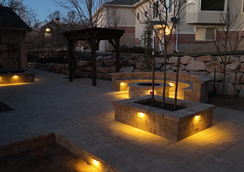 Outdoor patio hardscape featuring a stone fire pit, pergola, raised planters, and warm landscape lighting at dusk