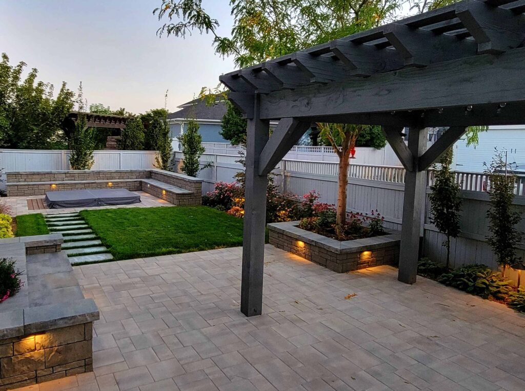 Modern backyard landscape design in Salt Lake City featuring a dark wood pergola, paver patio, raised planter beds, and integrated outdoor landscape lighting.