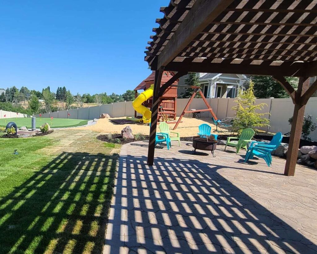 A West Jordan Utah family backyard featuring a shaded children's play area with rubber mulch, artificial turf dog zone, and patio seating for adults