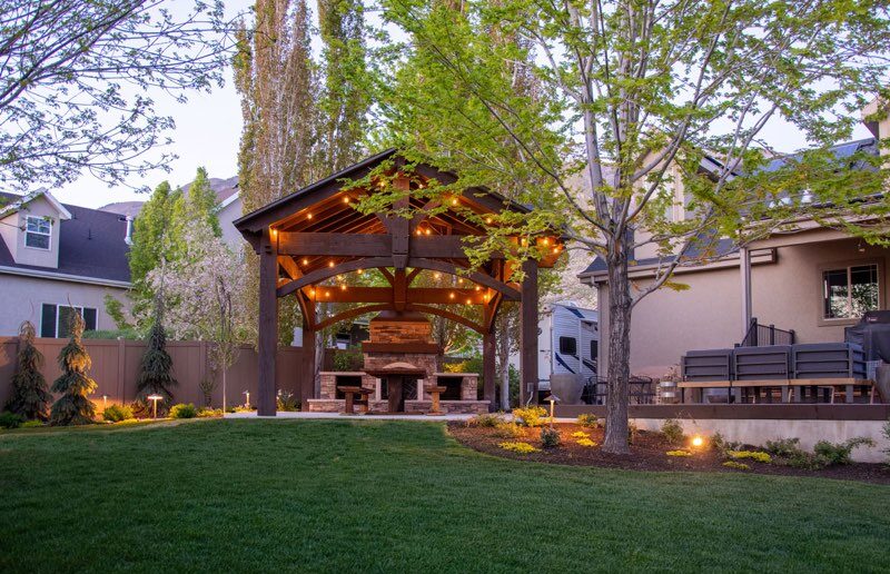 Custom timber pavilion with an outdoor stone fireplace, landscape lighting, mature trees, and a landscaped backyard.
